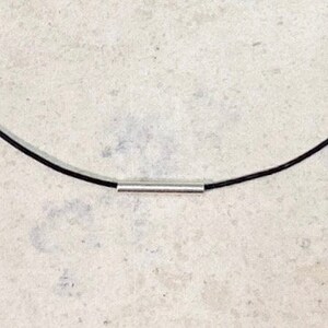 May include: A necklace featuring a thin, smooth black cord and a single, polished silver-toned cylindrical bead centered on the cord. The design is simple and consists of a continuous black line interrupted by the metallic silver tube component.