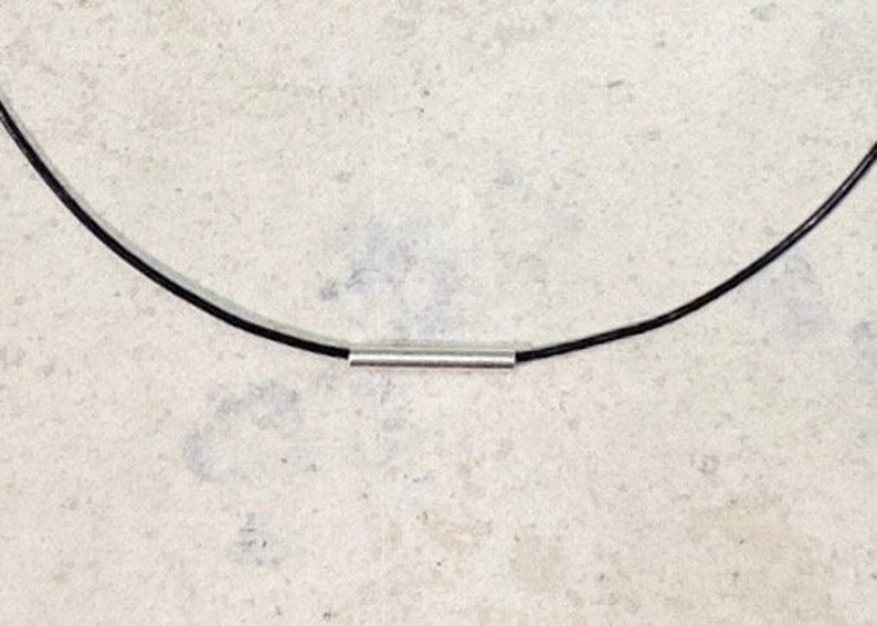 May include: A necklace featuring a thin, smooth black cord and a single, polished silver-toned cylindrical bead centered on the cord. The design is simple and consists of a continuous black line interrupted by the metallic silver tube component.