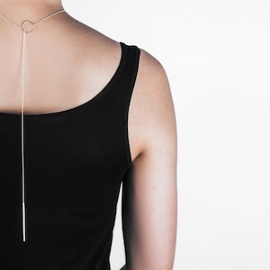 Puede incluir: Un collar largo y delgado de cadena plateada con un pequeño colgante circular. El collar lo lleva una persona que lleva una camiseta sin mangas negra.