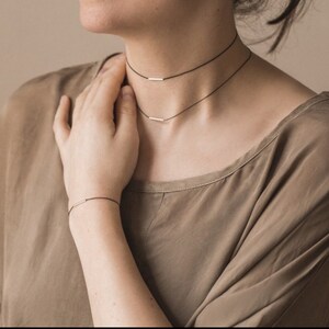 May include: A double-layered black cord choker necklace and a matching black cord bracelet, each featuring a single small, horizontal silver-toned metal bar accent. The jewelry set is worn around the neck and on the wrist, showcasing thin cords.