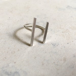 Peut inclure: Une bague ouverte de couleur argentée présentant deux barres rectangulaires verticales positionnées parallèlement l'une à l'autre. La bague possède une finition lisse et polie ainsi qu'un anneau fin et arrondi reliant les deux barres.