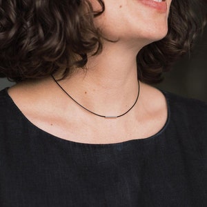 May include: A black cord necklace featuring a single, small, polished silver-toned metal bar pendant centered on the cord. The necklace rests against the neck, showcasing a simple design with a smooth, reflective finish on the metallic bar component.
