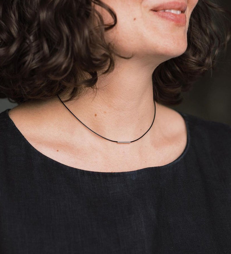 May include: A black cord necklace featuring a single, small, polished silver-toned metal bar pendant centered on the cord. The necklace rests against the neck, showcasing a simple design with a smooth, reflective finish on the metallic bar component.