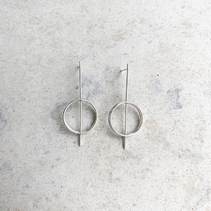 May include: A pair of silver-toned earrings featuring a thin vertical bar intersecting a circular hoop. The design consists of a straight metal post extending through the center of a round ring, creating a balanced geometric shape with a polished finish.