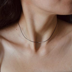 May include: A thin black cord choker necklace featuring a small, horizontal, polished silver-toned metal bar centered at the front. The necklace rests against the neck, providing a simple and clean aesthetic with its contrasting dark cord.