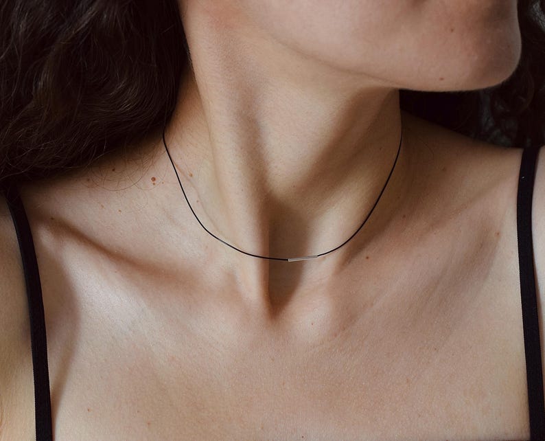 May include: A thin black cord choker necklace featuring a small, horizontal, polished silver-toned metal bar centered at the front. The necklace rests against the neck, providing a simple and clean aesthetic with its contrasting dark cord.