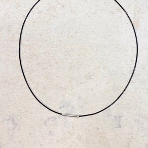 May include: A thin black circular necklace cord featuring a single small silver-toned cylindrical metal bead positioned at the bottom center. The necklace forms a simple, clean loop design with a smooth finish and a minimalist aesthetic structure.
