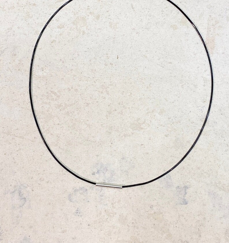 May include: A thin black circular necklace cord featuring a single small silver-toned cylindrical metal bead positioned at the bottom center. The necklace forms a simple, clean loop design with a smooth finish and a minimalist aesthetic structure.