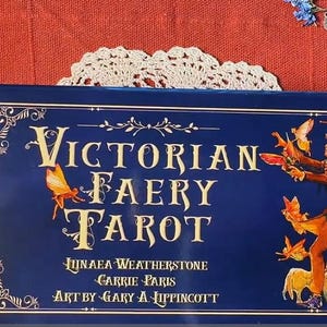 May include: A blue box with the title "Victorian Faery Tarot" in gold lettering. The box features illustrations of fairies and a whimsical design. The text "Juniaea Weatherstone, Carrie Paris, Art by Gary A. Lippincott" is printed on the bottom of the box.