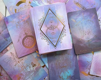 May include: A deck of tarot cards with a celestial theme. The cards feature watercolor backgrounds in shades of purple, blue, and pink, with gold line art illustrations. The deck is surrounded by other cards with various designs and titles.