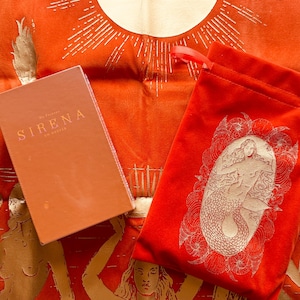 May include: A rectangular orange box with the text SIRENA printed in gold, placed next to a red drawstring pouch featuring a white line-art illustration of a mermaid, all resting on an orange fabric surface with a gold sun and mermaid print.