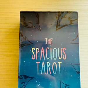 May include: A tarot card deck with a blue cover featuring the text "THE SPACIOUS TAROT" in colourful letters. The design includes illustrations of trees and a starry night sky.
