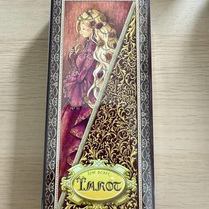 May include: A box for a tarot deck with a gold and burgundy design. The box features a woman with long blonde hair wearing a burgundy dress. The text "low scare TAROT" is on the front of the box.
