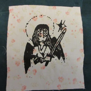 May include: A square fabric patch featuring a light cream background with a repeating pattern of small pink flowers. Centered is a black ink illustration of a multi-eyed figure with wings, a halo, and a sword, holding a star-shaped object.
