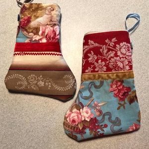 May include: Two decorative Christmas stockings. One features a dog and floral pattern, with red, brown, and cream colors. The other has a red floral pattern with a blue floral bottom. Both have hanging loops.