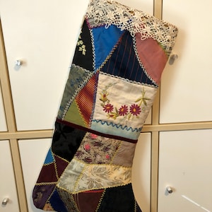May include: A patchwork Christmas stocking with a variety of fabrics in jewel tones and neutral colors. The stocking features embroidered flowers and a lace cuff. A white ribbon is attached for hanging.