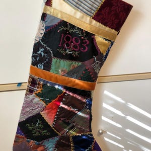 May include: A patchwork Christmas stocking with a variety of fabrics in jewel tones. The year "1983" is embroidered in pink thread. Gold ribbon and a white button add detail to this festive holiday decoration.