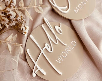 May include: Two beige round wooden signs with white lettering. One sign says "Hello World" and the other says "One Month".