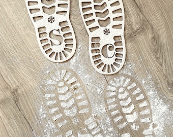 May include: Two wooden Christmas boot stencils with snowflake designs and the letters "S" and "C" cut out. The stencils are placed on a wood floor with white powder sprinkled around them, creating the impression of footprints.