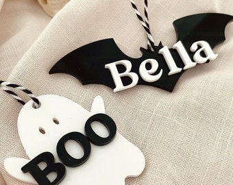 May include: Two Halloween-themed ornaments, one a white ghost with the word "BOO" in black letters, and the other a black bat with the name "Bella" in white letters.