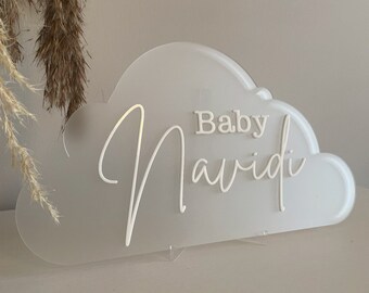 Może przedstawiać: Biały znak w kształcie chmury z napisem "Baby Navidi" w czcionce skryptowej. Znak wykonany jest z matowego materiału i jest wyświetlany na przezroczystym stojaku. Tło jest w neutralnym kolorze.