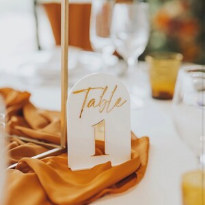 May include: White table number sign with gold lettering that says "Table 1" on a gold stand. The sign is on a table with a brown tablecloth.