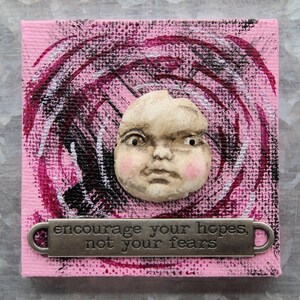 May include: A square canvas painted with a pink background and dark circular brushstrokes. A sculpted, pale, realistic face with rosy cheeks is centered. Below the face, a metal plaque features the text: encourage your hopes, not your fears.