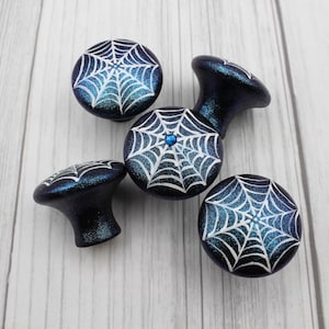 May include: A collection of dark blue cabinet knobs with a shimmering finish. Each knob features a white spiderweb design. One knob has a small blue gem in the center of the web. These decorative knobs are perfect for adding a spooky touch to furniture.