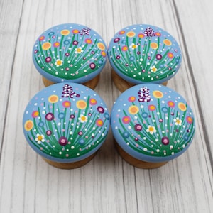May include: Four round, hand-painted cabinet knobs. Each knob features a light blue background with a floral design in yellow, pink, white, and green. The knobs are mounted on small wooden bases.