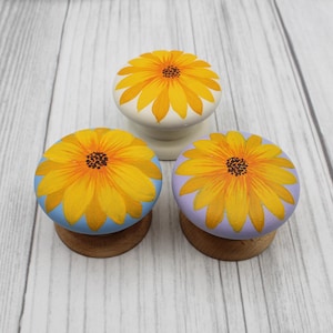 May include: Three decorative cabinet knobs with a sunflower design. The knobs feature a yellow sunflower with orange and brown details on a white, light blue, and lavender background. The knobs are mounted on wooden bases.