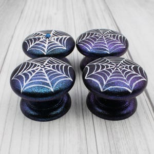May include: Four decorative cabinet knobs with a dark blue and purple glitter finish. Each knob features a white spiderweb design. One knob has a small blue gem. These knobs are perfect for adding a spooky touch to furniture.