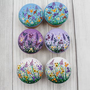 May include: Six round, hand-painted wooden bottle stoppers. The stoppers are painted with colorful floral designs in shades of blue, purple, yellow, and white. The stoppers have a cork base and are arranged on a light wood surface.