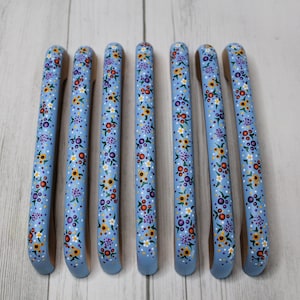 May include: Set of seven blue wooden hangers with a floral pattern painted on them. The flowers are in shades of yellow, orange, purple, and white.