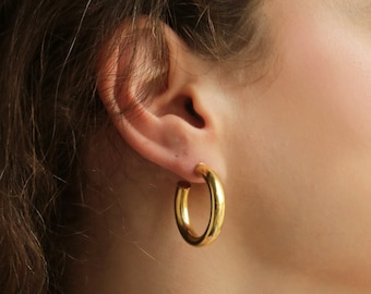 May include: A close-up of a person wearing a pair of gold hoop earrings. The earrings are thick and circular, reflecting light. The earrings are a bright, shiny gold color. The person's skin is fair, and their hair is brown.