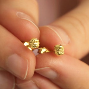 Könnte beinhalten: Ein Paar kleine, goldfarbene Ohrstecker. Jeder Ohrring hat ein strukturiertes, zylindrisches Design. Die Ohrringe werden zwischen zwei Fingern vor einem zartrosa Hintergrund gehalten. Die Ohrringe sind ein schlichtes, elegantes Schmuckstück.