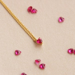 May include: A gold-coloured necklace featuring a heart-shaped, ruby-red pendant. Several loose, heart-shaped gemstones in a matching ruby-red colour are scattered around. The necklace has a delicate design, suitable for special events.