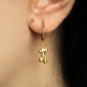 Puede incluir: Pendiente de aro dorado con un peque&ntilde;o colgante estilizado de torso femenino. El pendiente se lleva en la oreja, con el colgante colgando debajo del l&oacute;bulo. La joya es sencilla y elegante.