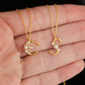 May include: Two gold-colored initial necklaces, one with the letter "C" and the other with the letter "S". Each pendant is adorned with small, clear crystals and a single pearl. The necklaces are displayed against a neutral background.