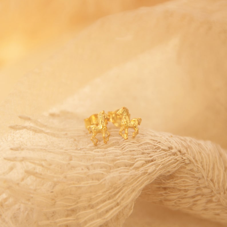 May include: A pair of gold-colored horse stud earrings. The earrings are small and detailed, with the horses in a running pose. The earrings are set against a soft, textured, beige background.
