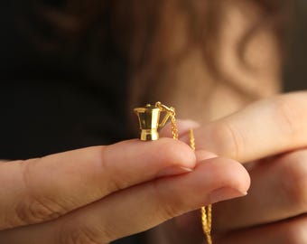 May include: A gold-colored pendant necklace featuring a miniature coffee pot charm. The charm is a detailed replica of a stovetop espresso maker. The necklace chain is also gold, creating a cohesive and elegant look. The necklace is held between fingers.
