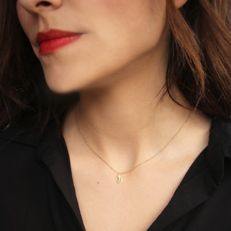 May include: A delicate gold-colored necklace with a small pendant. The pendant is a stylized figure. The necklace is worn against a black shirt. The person wearing the necklace has red lipstick.