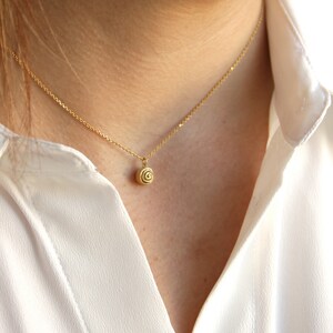 Op de afbeelding: Een delicate gouden ketting met een kleine, spiraalvormige hanger. De hanger hangt aan een fijne ketting. De ketting wordt gedragen over een wit overhemd. Een eenvoudig, elegant sieraad.
