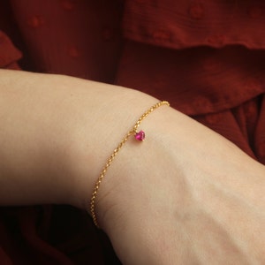 Peut inclure: Un bracelet en or délicat avec un petit charme en forme de cœur en pierre précieuse rouge. Le bracelet présente un design de chaîne fine et est présenté sur le poignet d'une personne sur un fond rouge. Le charme est d'un rouge vif.