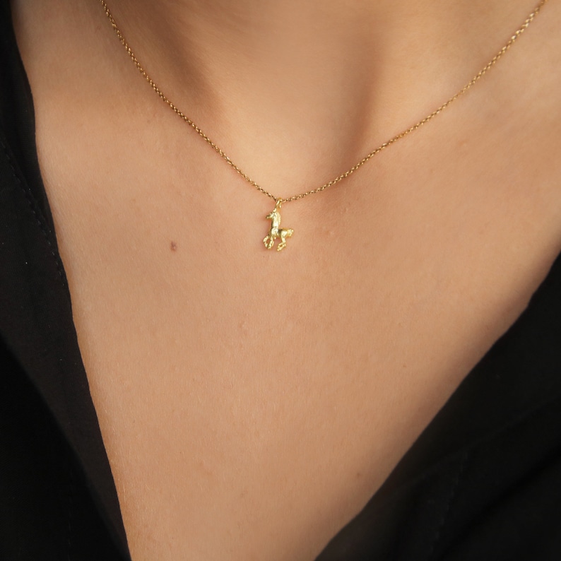 May include: A delicate gold-colored necklace featuring a small, textured horse pendant. The horse charm is suspended from a fine chain, creating a minimalist and elegant look. The necklace is shown against a dark background.