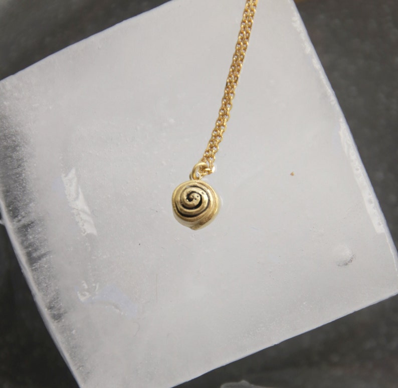 Op de afbeelding: Een goudkleurige ketting met een kleine, spiraalvormige hanger. De hanger hangt aan een fijne ketting. De ketting wordt gepresenteerd tegen een lichtgekleurde, getextureerde achtergrond.