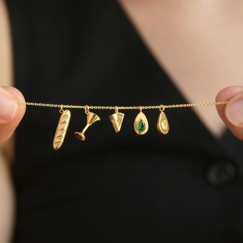 May include: A gold necklace featuring charms shaped like a baguette, cocktail glass, cheese wedge, avocado with a green stone, and avocado half. The charms are suspended from a delicate gold chain, creating a unique and stylish accessory.