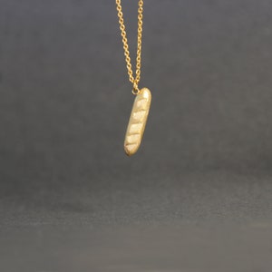May include: A gold-colored pendant necklace featuring a baguette-shaped charm. The charm has a textured surface with horizontal lines, resembling a loaf of bread. The necklace chain is also gold-colored, and the background is a dark gray.