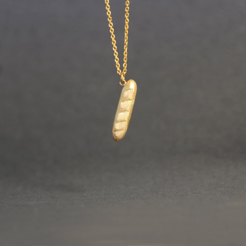 May include: A gold-colored pendant necklace featuring a baguette-shaped charm. The charm has a textured surface with horizontal lines, resembling a loaf of bread. The necklace chain is also gold-colored, and the background is a dark gray.