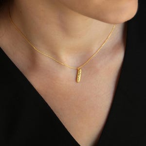 May include: A gold-colored necklace with a small, textured, vertical pendant. The pendant has a series of small, raised details. The necklace is worn against a dark background.