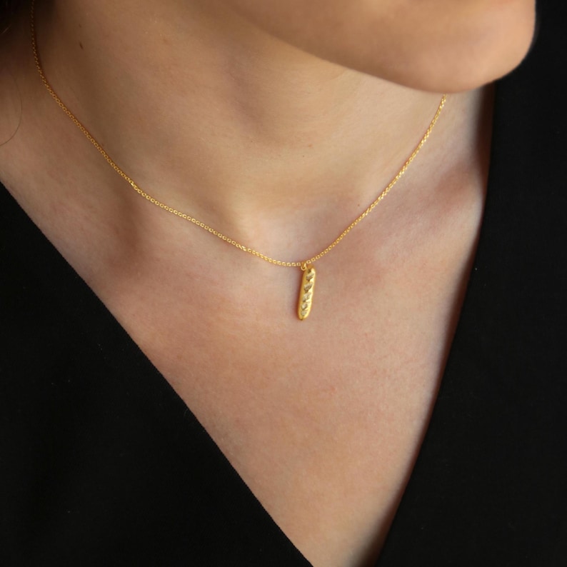 May include: A gold-colored necklace with a small, textured, vertical pendant. The pendant has a series of small, raised details. The necklace is worn against a dark background.